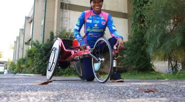 vanessa está ajoelhada no chão ao lado da bicicleta adaptada. #paratodosverem