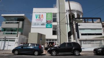 Fachada de vila criativa com placa no alto e carros estacionados na porta. #paratodosverem