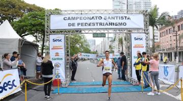 Homem cruza a linha de chegada onde há um portal onde de se lê Campeonato Santista de Pedestrianismo