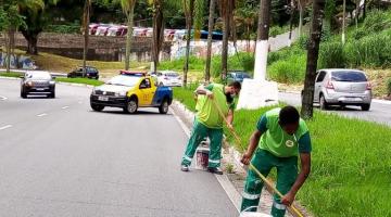 dois homens estão caiando guia junto a canteiro central. Eles seguram cabos e têm à frente latas com tinta. Ao fundo, um veículo da CET sinaliza a via. #paratodosverem