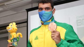 O atleta segura o boneco do mascote da competição na mão direita e a medalha na esquerda. Ele está de uniforme e máscara. #paratodosverem