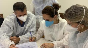 agentes de saúde paramentados estão mexendo em papeis com anotações em cima de uma mesa. #Paratodosverem