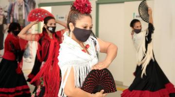 close de mulher dançando flamenco, com roupa típica, o que inclui xale nos ombros, saia rodada e flor na cabeça, além de um leque aberto nas mãos. Atrás há outras mulheres também dançando. #paratodosverem