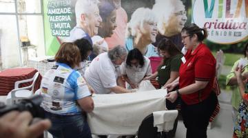 Mulheres em pé em volta de tecido aprendendo sobre o bordado. #pratodosverem