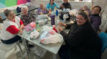 mulheres estão reunidas em torno de mesa fazendo artesanato. Algumas olham para a foto e sorriem. #paratodosverem 