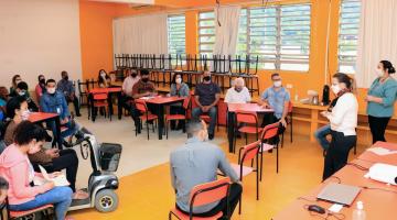 Sala de aula com cadeiras dispostas em círculo. Pessoas estão sentadas distantes umas das outras. #paratodosverem