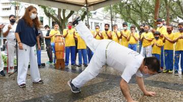 homem está jogando capoeira com as duas mãos e um dos pés no chão. A outra perna está no ar. Ele está de costas. O público aplaude ao fundo. #paratodosverem