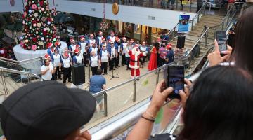 integrantes de coral estão em formação de apresentação com uma grande árvore de natal ao fundo. Em primeiro plano, o público tira fotos da apresentação com celulares. #paratodosverem