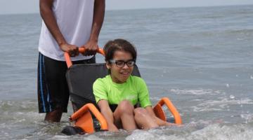 Criança toma banho de mar com ajuda de cadeira anfíbia e monitor. #pracegover