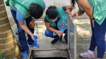 Agentes vistoriam caixa d'água usando lanternas. São três pessoas usando coletes de identificação. #Paratodosverem