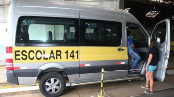 van escolar está parada em baia. Há uma pessoa sentada no banco do passageiro olhando o interior do veículo. Outra, está encostada na porta observando. #paratodosverem