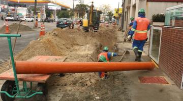 homens trabalhando em calçada aberta. Há grande volume de terra e areia em torno além de um cano em primeiro plano. #paratodosverem