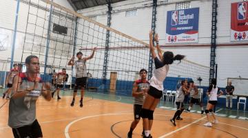 jovens estão na quadra jogando vôlei. A rede aparece no meio com alunos de cada um dos lados. #paratodosverem