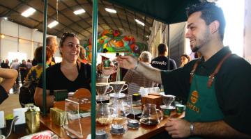 homem serve café para mulher em estande com vários tipos de café #paratodosverem