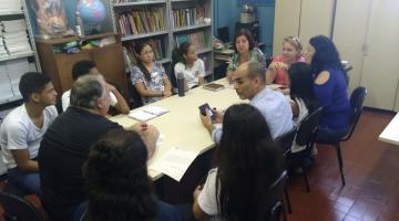 Representantes de secretarias, da escola e alunos estão sentados em torno de uma mesa. Em uma das paredes está encostada uma estante de livros. #Pracegover