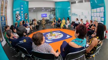 Várias pessoas estão reunidas em um círculo. Há uma mesa ao centro com o bastão que será passado entre os integrantes do círculo. #Pracegover