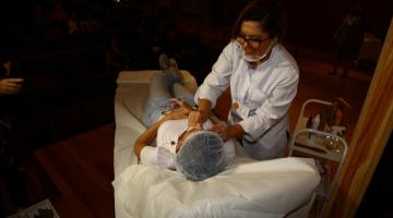 Esteticista está em pé atendendo uma pessoa que está deitada em maca. A profissional passa as mãos no rosto da mulher deitada. #Pracegover