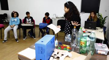 Crianças estão em uma sala. Uma delas está em pé ao lado de vários produtos recicláveis, como garrafas PET, rolos e papel e outros objetos. #Pracegover