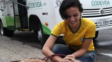 Mulher está agachada e passa a mão na cabeça de um cachorro. Ao fundo está o castramóvel. #Pracegover