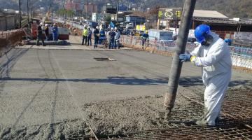 Homem com uniforme branco e capacete azul manuseia mangueira que despeja concreto sobre pista de viaduto