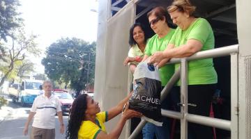 Com camisa da seleção brasileira de futebol, mulher entrega sacola com roupas para três mulheres de verde sobre caminhão