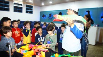 professor está com peixe de brinquedo na mão, observado por crianças #pracegover 