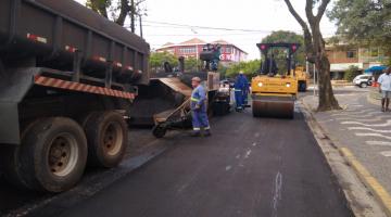 Rua é pavimentada. Caminhões e máquina compactadora estão na via. Homens também trabalham no local. #Pracegover