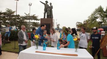 Celebração pela data no emissário submarino. Ao fundo está o monumento em homenagem à imigração japonesa no Brasil e, à frente uma mesa com vasos de flores e pessoas se cumprimentando. #Pracegover