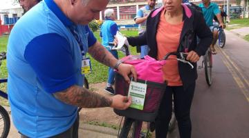 Agente da CET coloca placa em biclicleta. Uma mulher está desmontada da bike aguardando a afixação. Atrás há movimento de bicicletas e outros agentes. #Pracegover