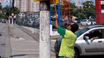 Homem remove banner afixado em poste. Ele usa uma vara para alcançar a propaganda. #Pracegover