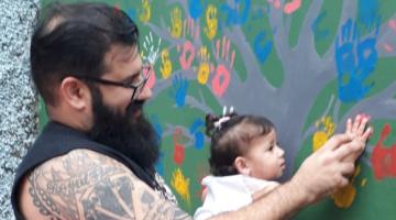homem encosta mão da filha em parede pintada de flores #pracegover 