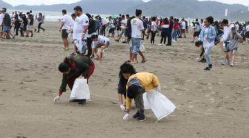 Três pessoas agachadas recolhem lixo da areia da praia, com cerca de 100 pessoas ao fundo vom sacolas plásticas nas mãos