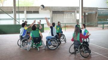 Seis jovens em cadeira de rodas erguem as mãos em disputa de bola de basquete dentro de quadra