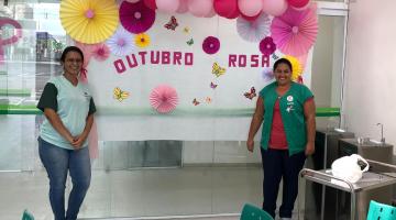 Funcionárias de policlínica estão diante de um painel com balões de gás cor de rosa. Ao fundo se lê Outubro Rosa. #Pracegover