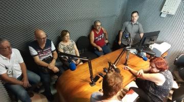 grupo está sentado na mesa da rádio com microfones no meio #pracegover 