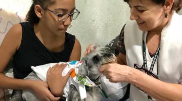 Menina segura cão em seu colo. Ao lado dela está uma mulher que usa um avental branco. #Pracegover