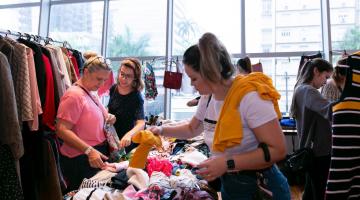 Bazar com mesa repleta de peças de roupas. Há mulheres examinando as peças. À esquerda há uma arara com várias roupas penduradas. #Pracegover