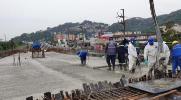 Vista geral de pista concretada, com homens uniformizados trabalhando. Em primeiro plano há umas ferragems aparentes. #Pracegover