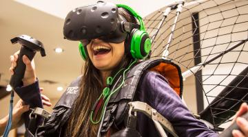 Menina está em um simulador de paramotor e tem nos olhos um óculos de realidade virtual. #Pracegover