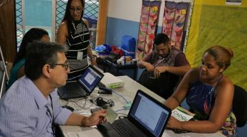 #pracegover Sentado à mesa, homem e mulher com notebooks cadastram homem e mulher sentados do lado oposto