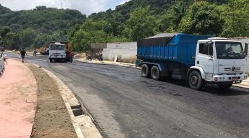 Vista geral de pista alfaltada. Ao lado direito se vê dois caminhões. Do esquerdo, o trecho de uma ciclovia. #paratodosverem