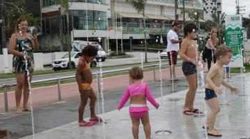 #pracegover Abservadas por três adultos, duas meninas e dois meninos brincam sobre a fonte interativa