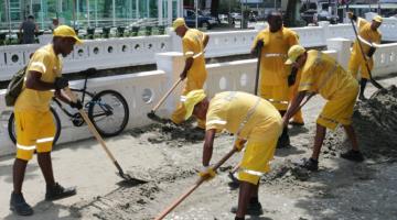 #pracegover Junto a mureta de canal na orla da praia, seis homens uniformizados de amarelo removem areia com pás