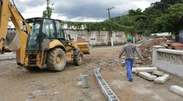 #pracegover homem com capacete caminha ao lado de escavadeira em terreno com materiais de construção junto à avenida