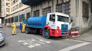 Caminhão com hidrojato está parado em rua ao lado de imóvel. Homens estão manuseando máquina na parte de trás do veículo. #Paratodosverem