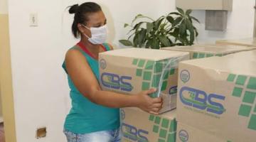Mulher usando máscara pega uma caixa de mantimentos de pilha de caixas. #Paratodosverem