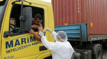 Pessoa uniformizada de branco, com avantal, touca, máscara e luva, em pé, vacina caminhoneiro que está dentro do veículo com o braço para fora. O caminhão é uma carreta com um contêiner no reboque. #Paratodosverem