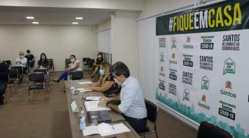 Sala com um painel à direita na parede onde se lê Fique em Casa e vários logotipos municipais abaixo. Em uma mesa estão sentadas duas pessoas usando máscaras. Ao fundo há quatro pessoas sentadas em várias cadeiras disponíveis. #Paratodosverem