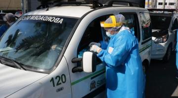 #pracegover com capa azul e proteção facial, enfermeira aplica teste em motorista pela janela do veículo sob tenda