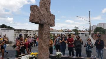 Pessoas rezando em direção a monumento de uma cruz no cemitério #paratodosverem